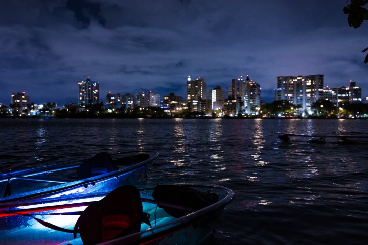 City skyline at night with illuminated boats on a calm river in the foreground.