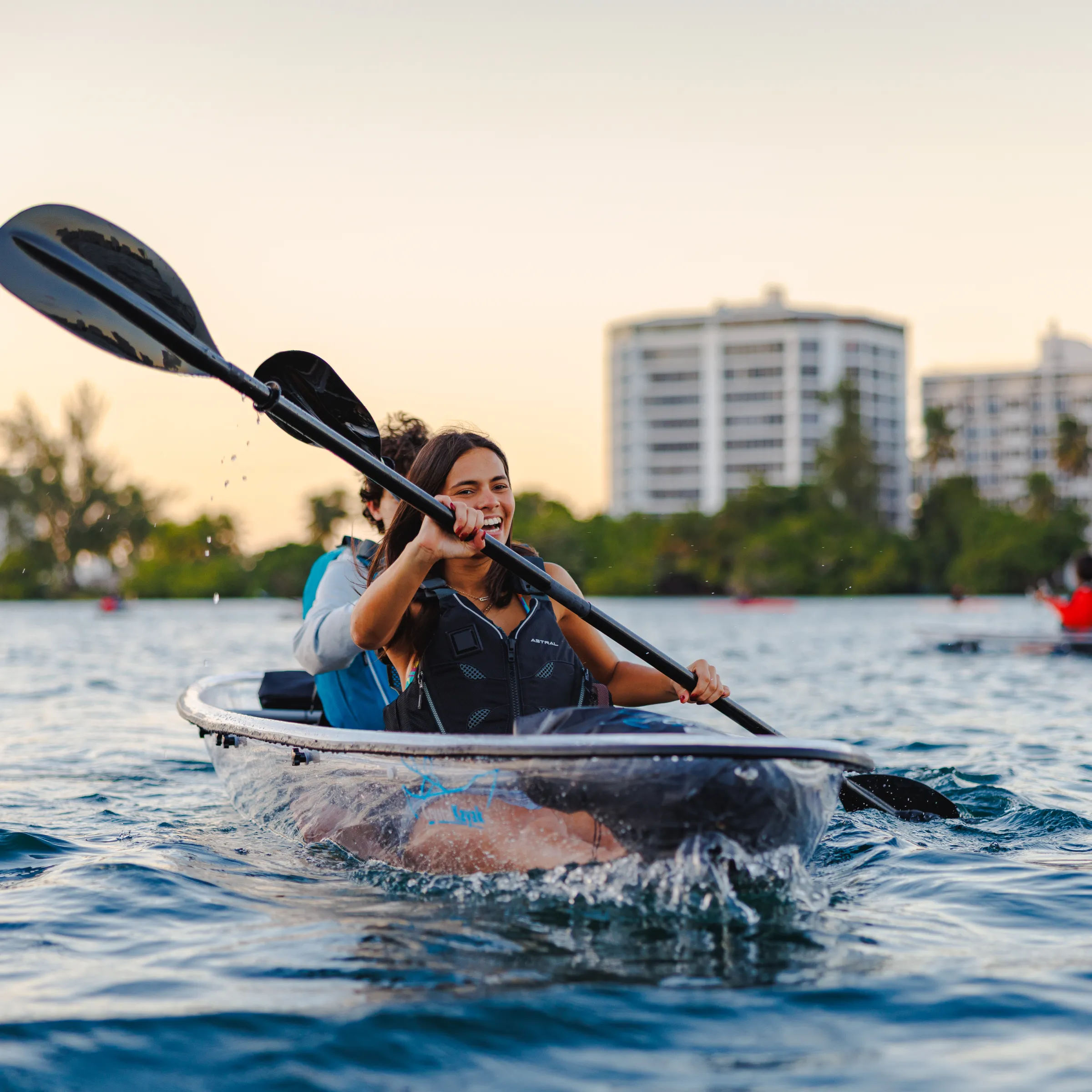 People kayaking on a body of water, with city buildings in the background during sunset.