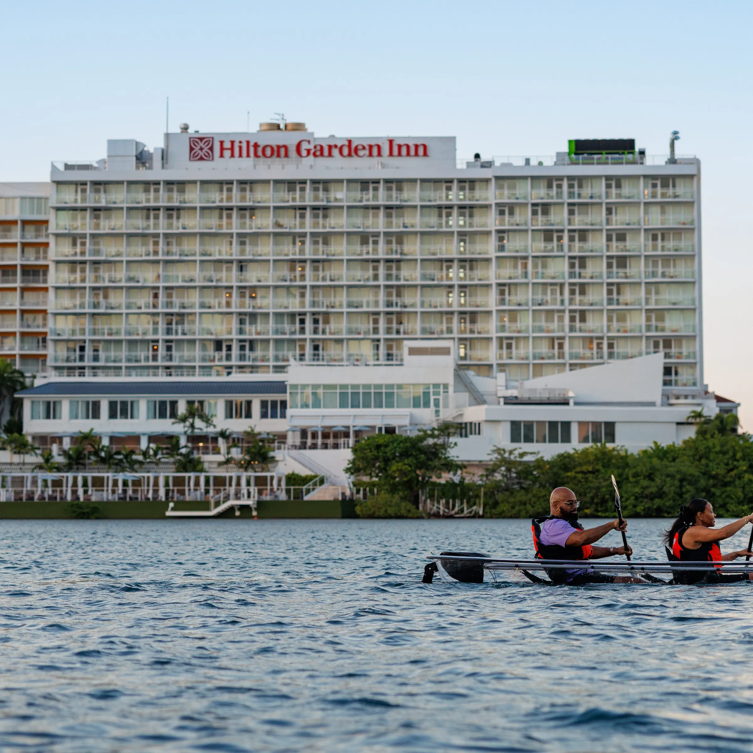 Two people kayaking on water near a Hilton Garden Inn hotel at sunset.