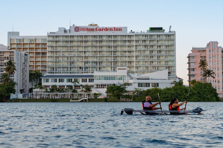 Two people kayaking on water near a Hilton Garden Inn hotel at sunset.