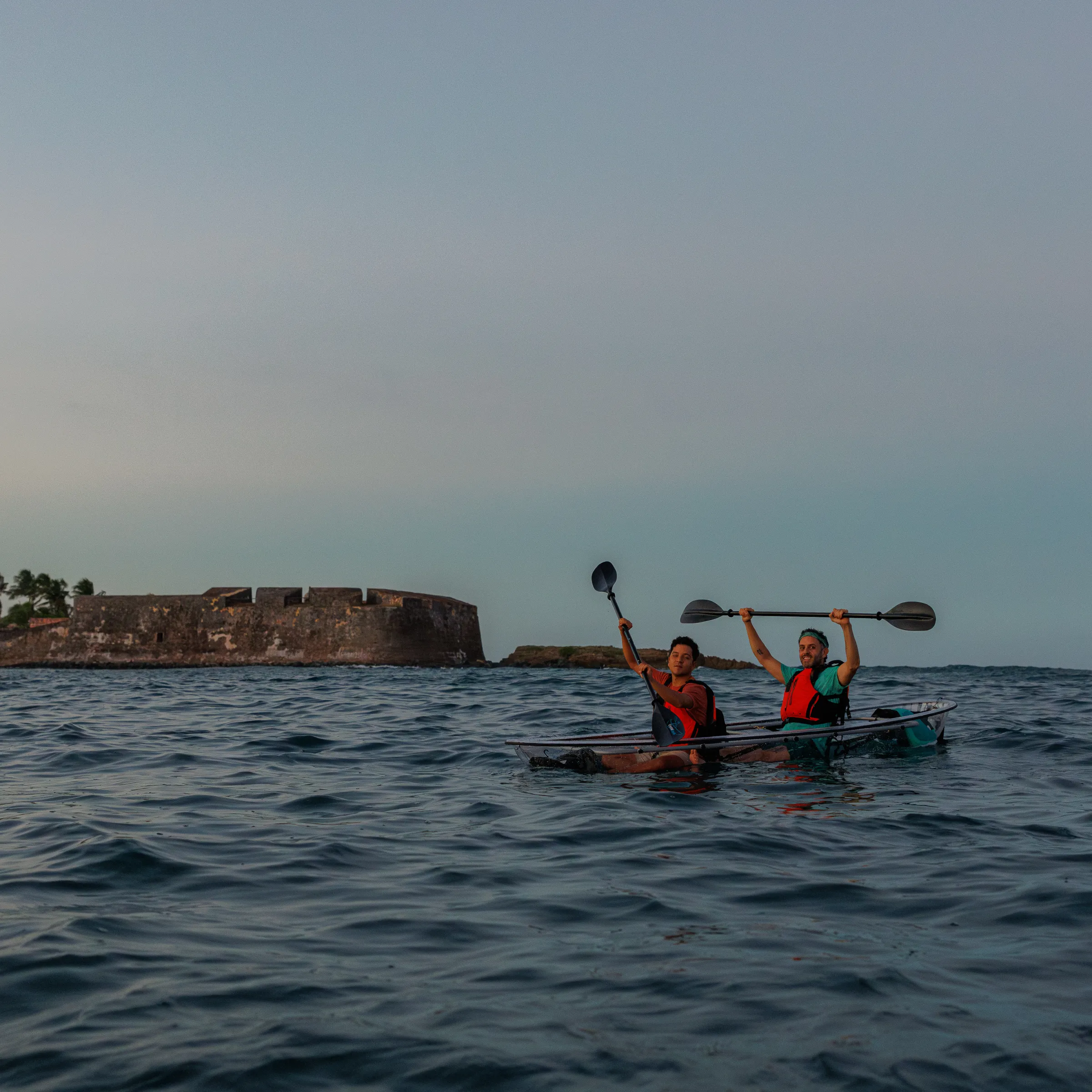 Two people kayaking near a stone fortress and palm trees in the ocean at sunset.
