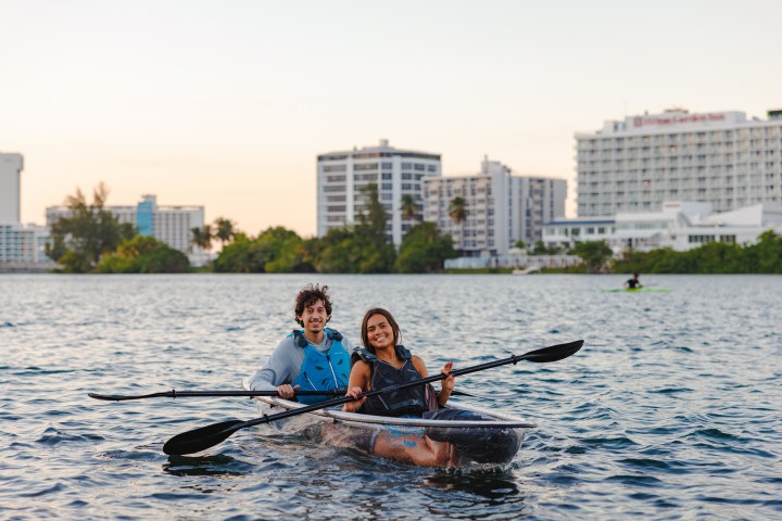 Two people kayaking on a lake with city buildings in the background.