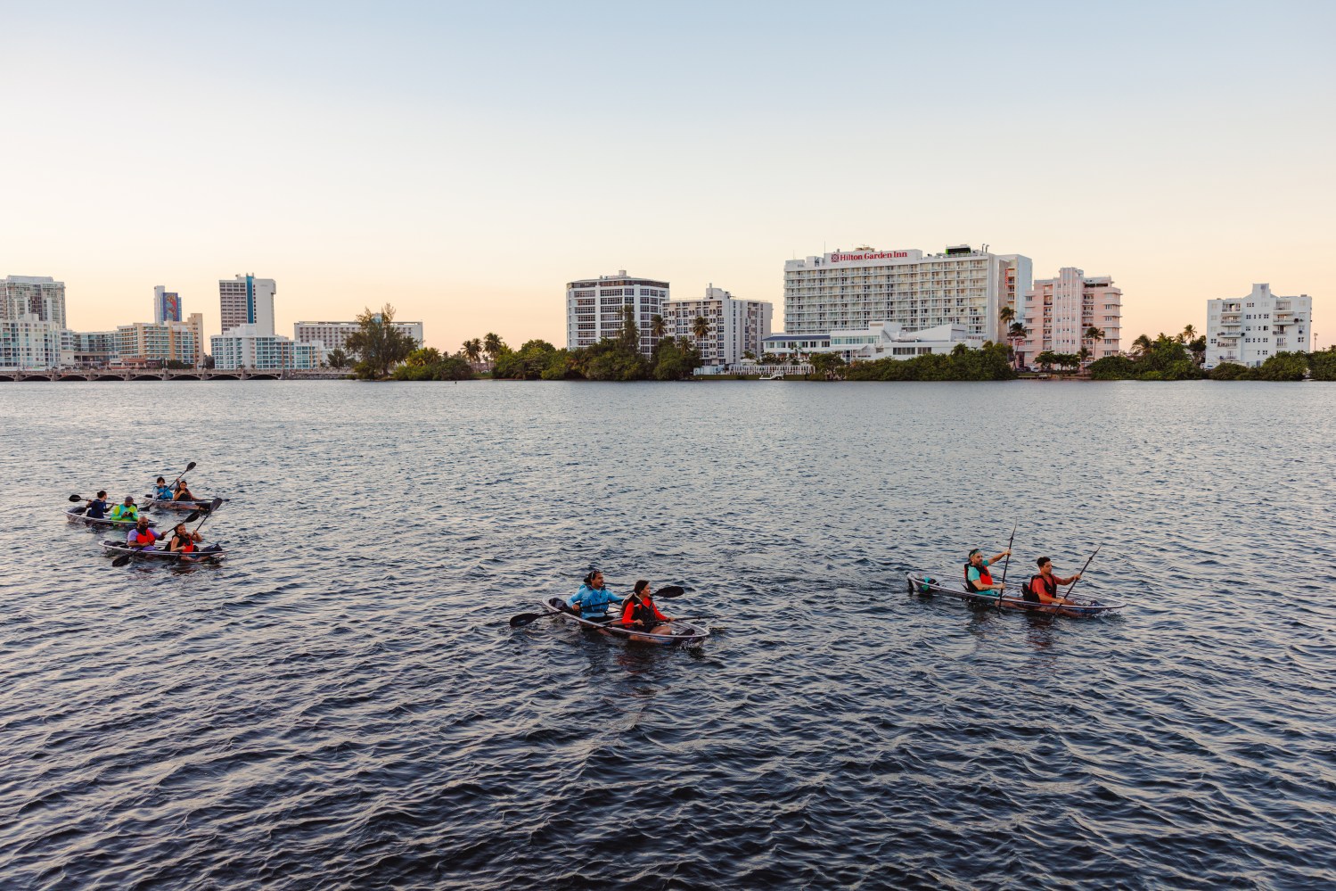 People kayaking on a wide river with city buildings in the background during sunset.
