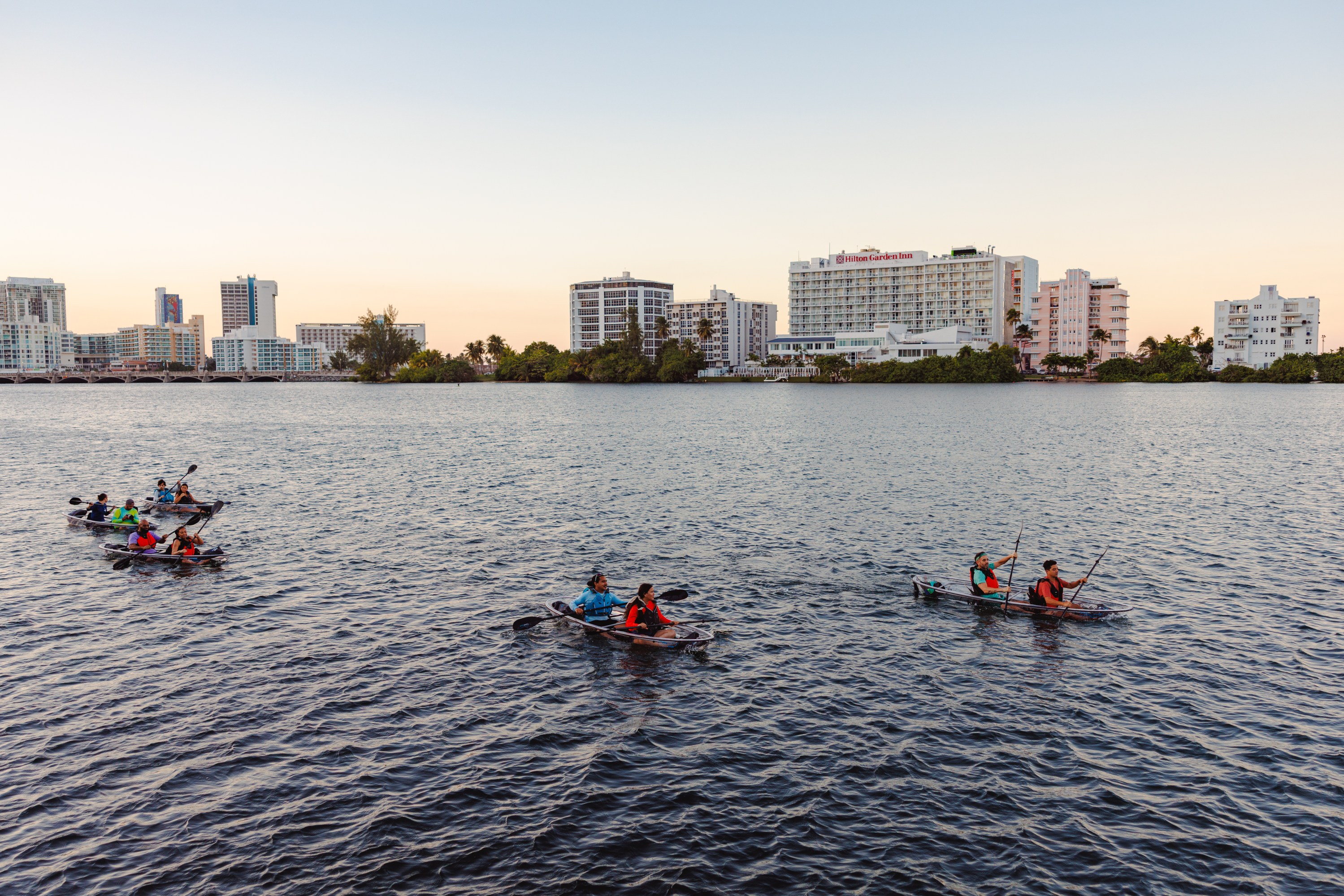 People kayaking on a wide river with city buildings in the background during sunset.