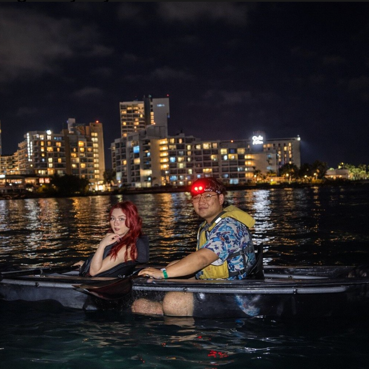 Two people in a kayak on water at night with city lights in background.