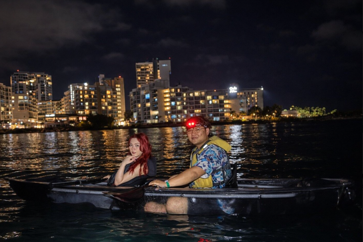 Two people in a kayak on water at night with city lights in background.