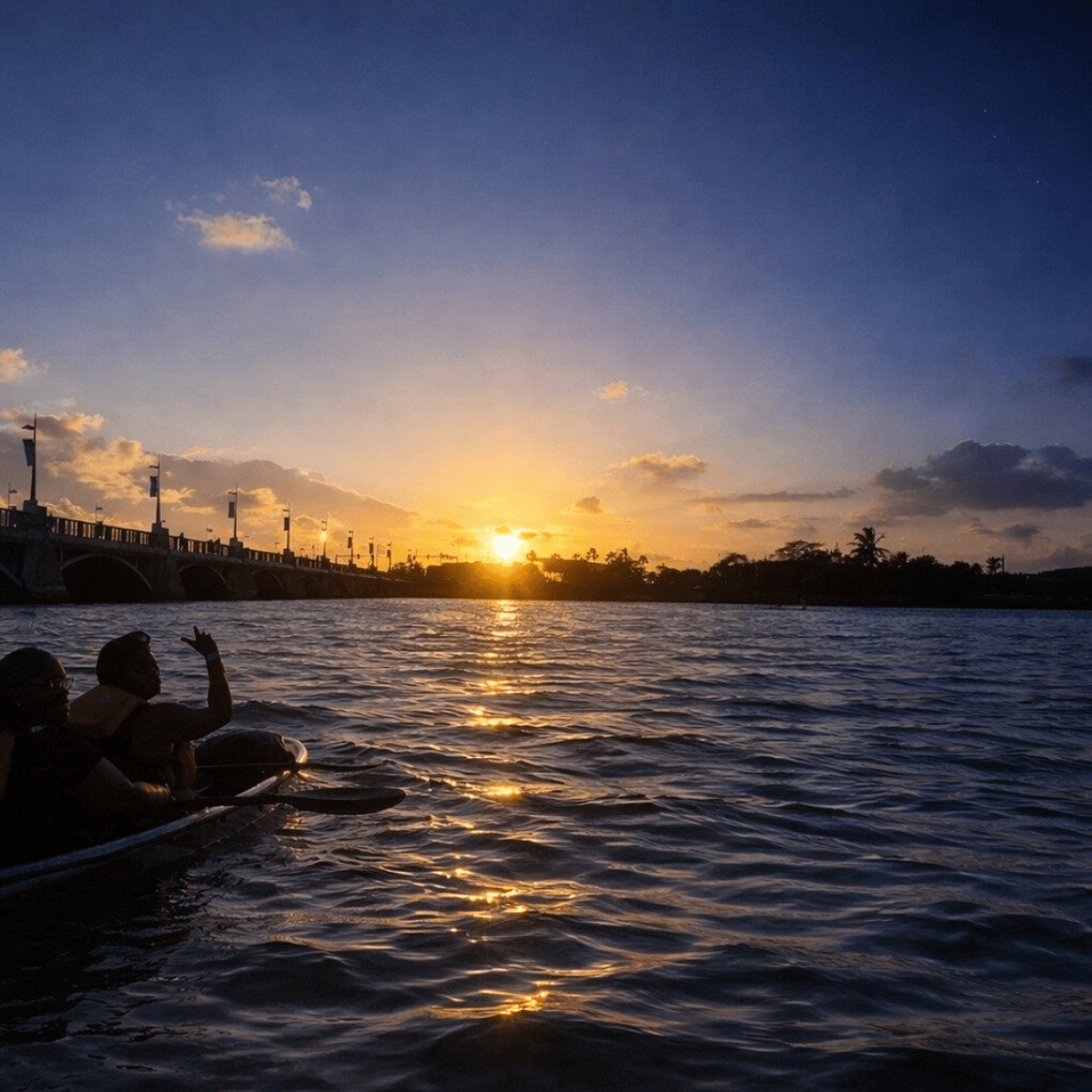Two people kayaking on a river at sunset under a bridge.