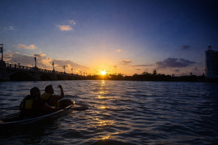 Two people kayaking on a river at sunset under a bridge.