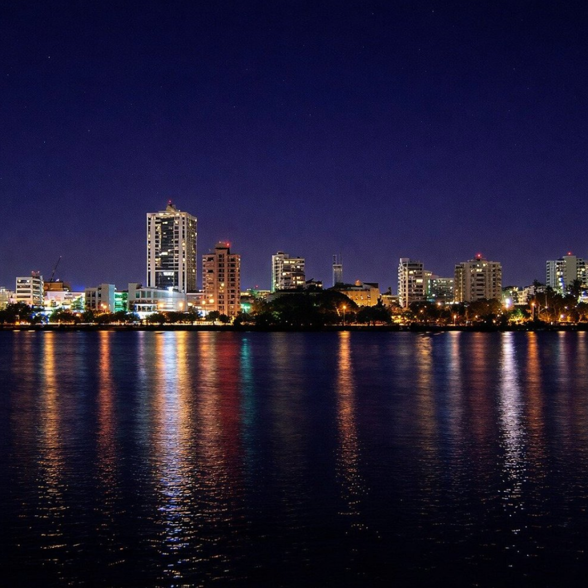 City skyline at night with buildings reflecting on a calm body of water.
