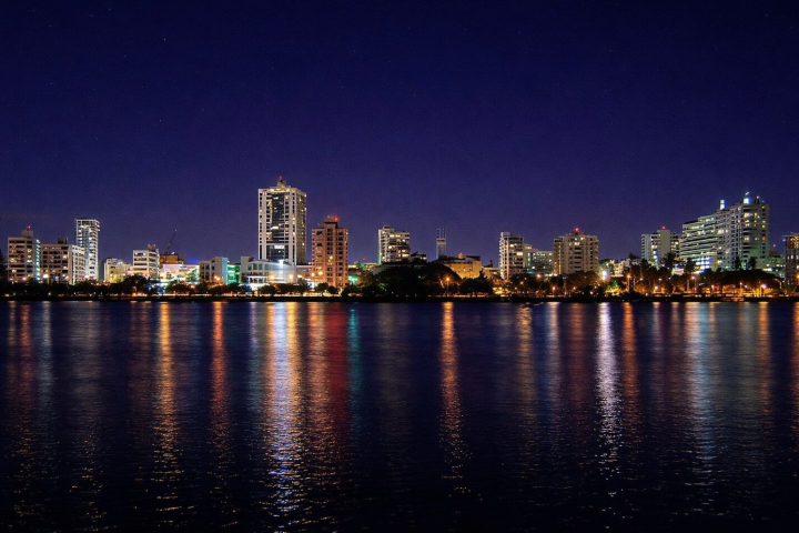 City skyline at night with buildings reflecting on a calm body of water.