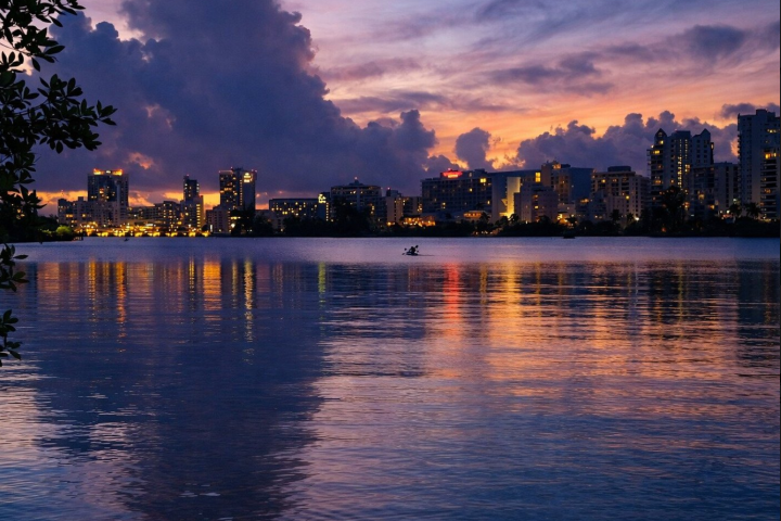 City skyline at dusk with reflections on the water and a cloudy sky.